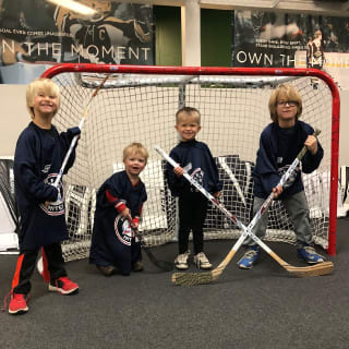 Four Young Kids Holding Hockey Sticks and Posing For Picture In Front Of Outdoor Street Hockey Net 